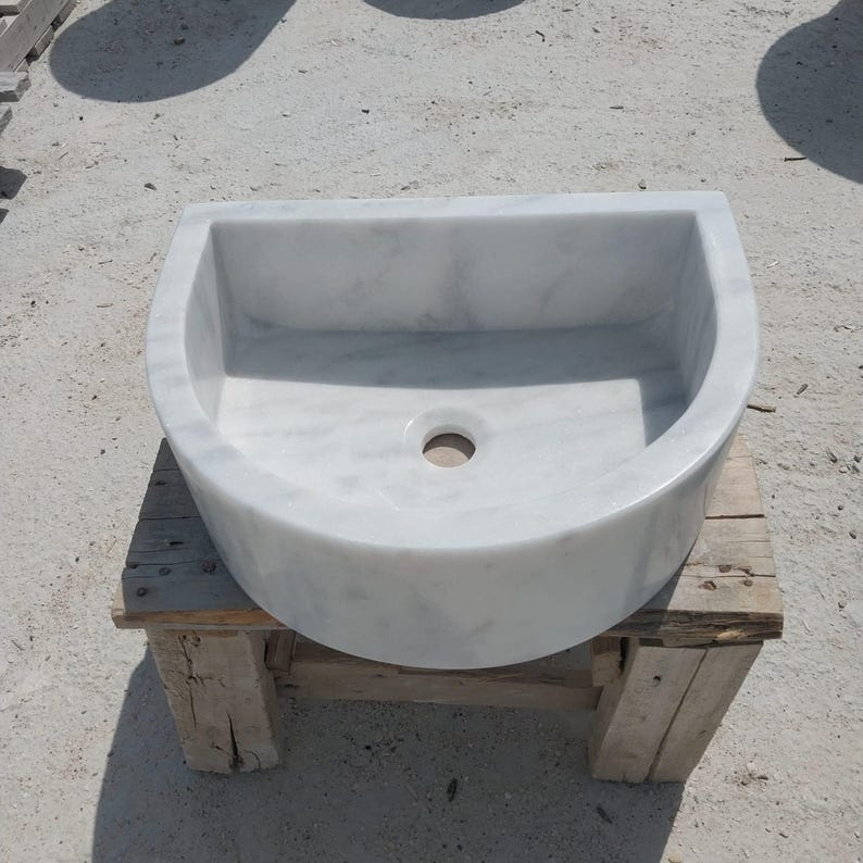 May include: A white marble sink with a semi-circular design. The sink has a smooth, polished surface and a central drain hole. It is placed on a rustic wooden stand, showcasing its elegant form.