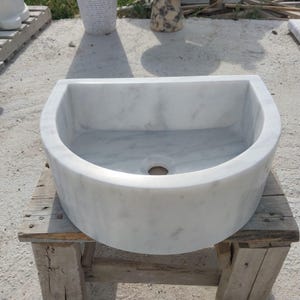 May include: A white marble sink with a semi-circular design. The sink has a smooth, polished surface and a drain hole in the centre. It is placed on a rustic wooden stand, showcasing its natural stone texture.