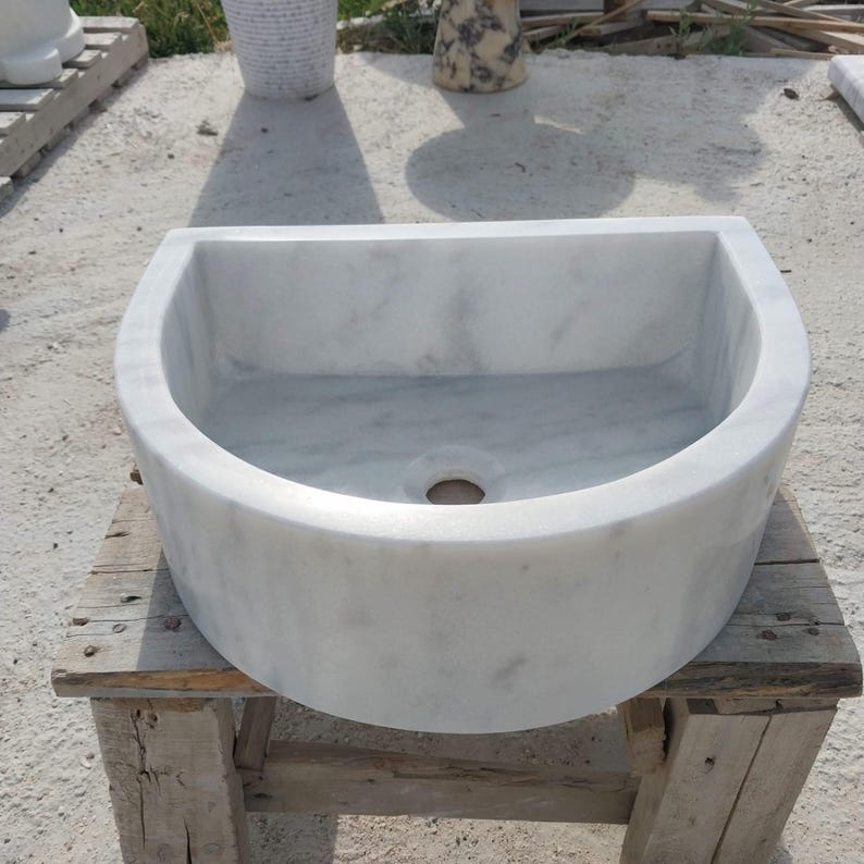 May include: A white marble sink with a semi-circular design. The sink has a smooth, polished surface and a drain hole in the centre. It is placed on a rustic wooden stand, showcasing its natural stone texture.
