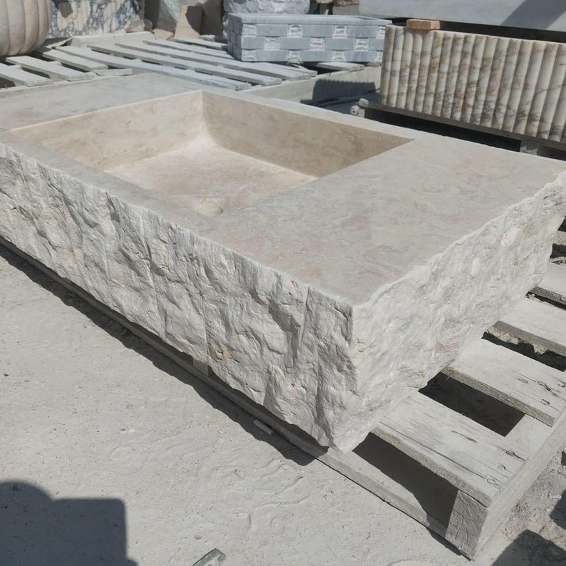 May include: A rectangular stone sink featuring a rough, chiseled exterior texture and a smooth, polished interior basin. The sink is crafted from light beige natural stone and displays a drain hole at the bottom of the basin, resting on a wooden pallet.