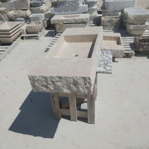 May include: A rectangular beige stone sink featuring a chiseled, rough-hewn exterior edge and a smooth, polished interior basin with a central drain hole. The sink is supported by a simple wooden frame structure, showcasing natural stone texture.