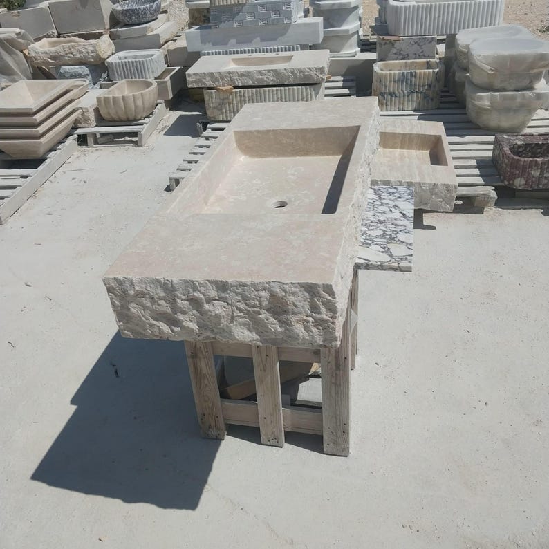 May include: A rectangular beige stone sink featuring a chiseled, rough-hewn exterior edge and a smooth, polished interior basin with a central drain hole. The sink is supported by a simple wooden frame structure, showcasing natural stone texture.
