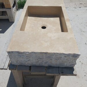 May include: A rectangular beige stone sink featuring a smooth, flat top surface with a central drain hole and a rough, chiseled, natural rock texture along the front edge. The basin is recessed into the stone block, showcasing a light tan color.
