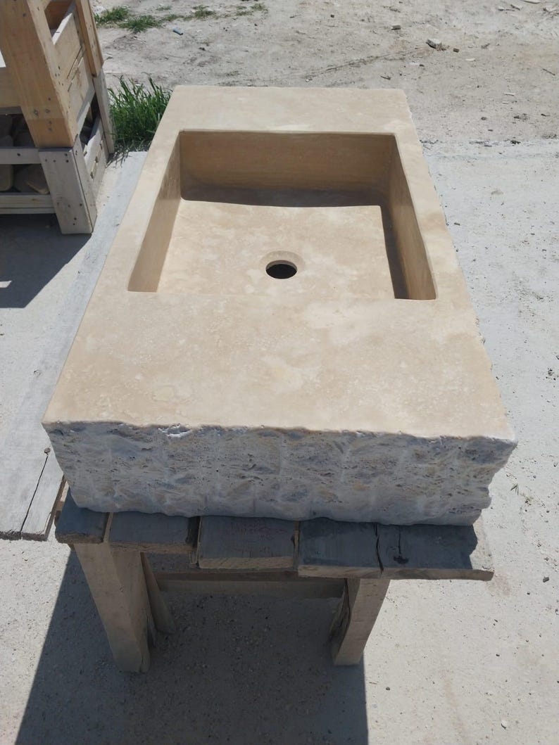 May include: A rectangular beige stone sink featuring a smooth, flat top surface with a central drain hole and a rough, chiseled, natural rock texture along the front edge. The basin is recessed into the stone block, showcasing a light tan color.