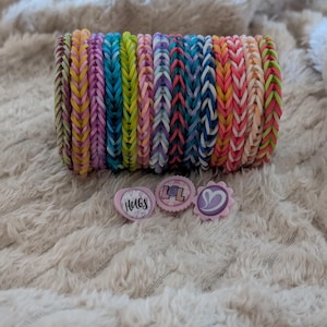 May include: A stack of colorful rubber band bracelets in various shades of pink, blue, green, and yellow. Three small, pink, decorative pins with the words "HUGS" and "LOL" and a heart design are also visible.