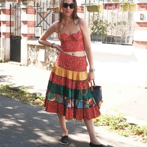 May include: A woman in a red floral crop top and a tiered midi skirt with red, yellow, and green panels. The skirt features a patchwork design on the bottom tier. She wears black flats and sunglasses, carrying a navy tote bag.