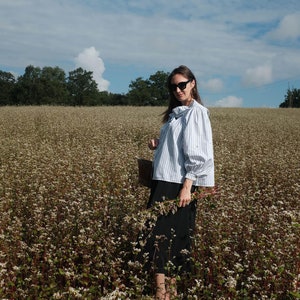 May include: A woman wearing a blue and white striped shirt with a bow tie, black skirt, and sunglasses stands in a field of white buckwheat flowers.