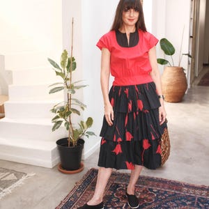 Könnte beinhalten: Ein rotes Oberteil mit schwarzem Ausschnitt und kurzen Ärmeln, kombiniert mit einem schwarzen Stufenrock mit rotem Blumenmuster. Das Outfit wird mit schwarzen Ballerinas getragen. Der Hintergrund ist ein Innenraum mit Teppich und Pflanzen.