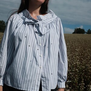 May include: A woman wearing a light blue and white striped button-up shirt with a bow tie. The shirt has a ruffled collar and long sleeves. She is standing in a field of white flowers.
