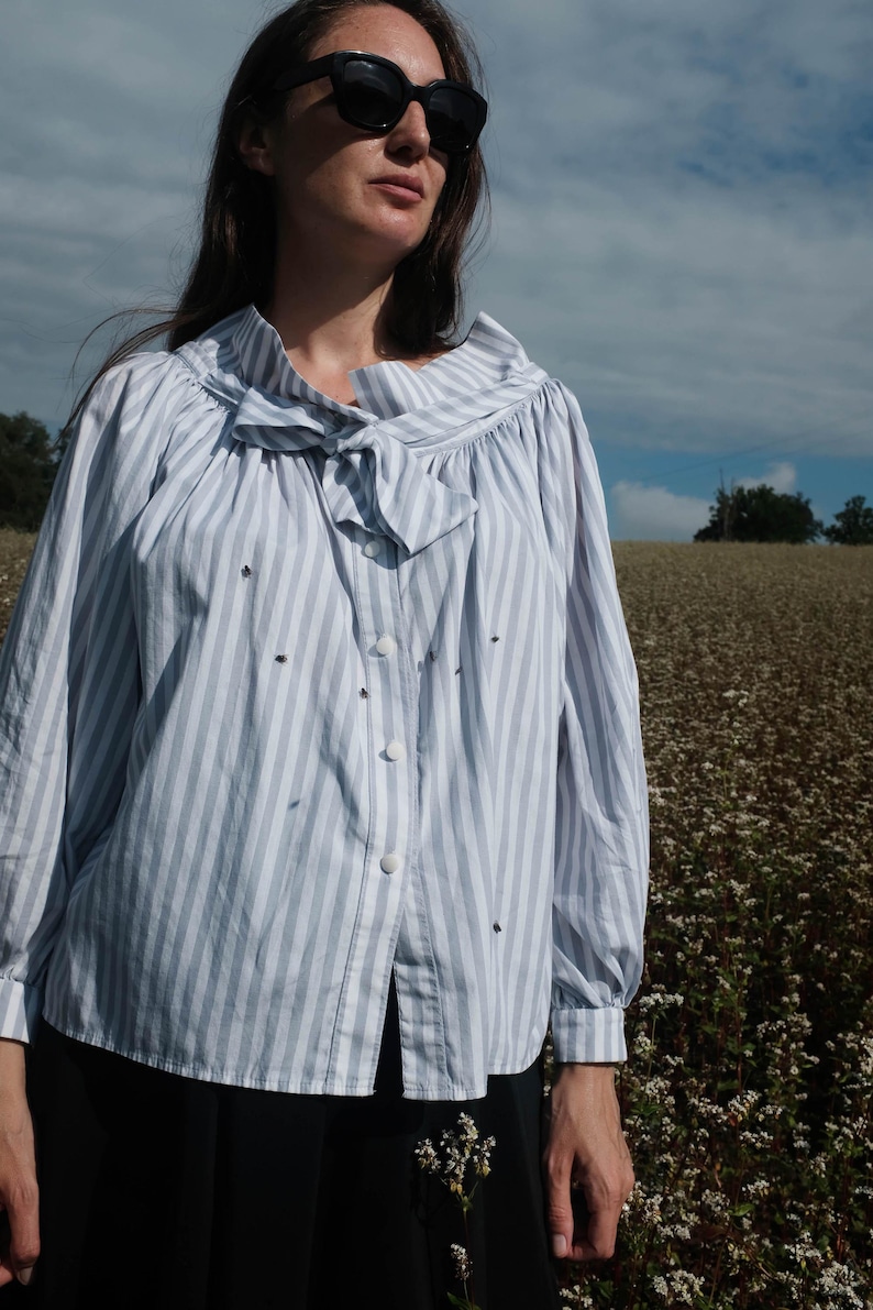 May include: A woman wearing a light blue and white striped button-up shirt with a bow tie. The shirt has a ruffled collar and long sleeves. She is standing in a field of white flowers.
