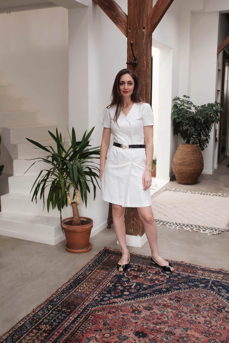 May include: A white dress with a black belt and a V-neckline. The dress is short-sleeved and has a wrap-style design. The woman is standing on a patterned rug.