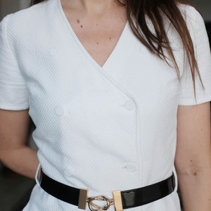 May include: A white dress with a V-neckline and short sleeves. The dress has a black belt with a gold buckle.