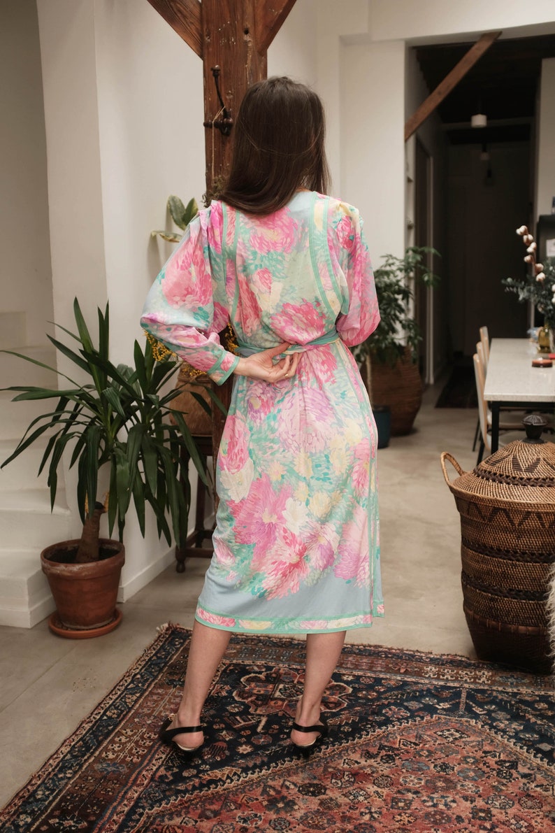 May include: A long, floral print dress with a light blue background and pink, yellow, and green flowers. The dress has a cinched waist and long sleeves. The model is wearing black heels.
