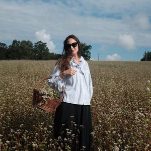 May include: A woman wearing sunglasses and a striped blue and white shirt stands in a field of white flowers. She is holding a wicker basket with flowers in it.
