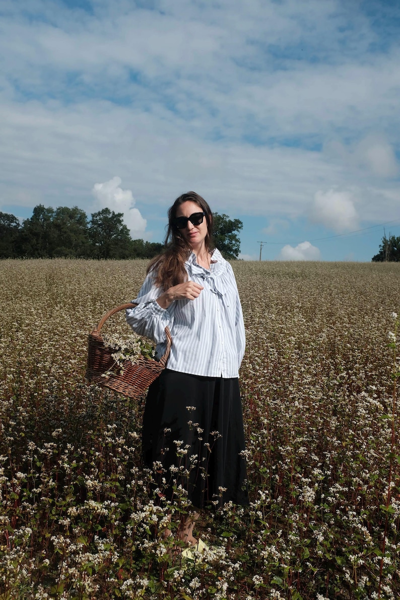 May include: A woman wearing sunglasses and a striped blue and white shirt stands in a field of white flowers. She is holding a wicker basket with flowers in it.