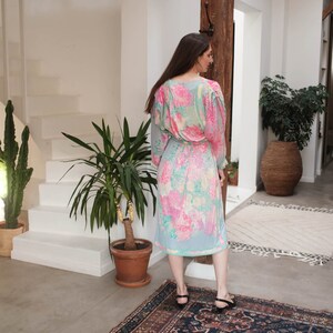 May include: A woman wearing a light blue midi dress with a pink and yellow floral print. The dress has a V-neckline and long sleeves. She is standing on a patterned rug.