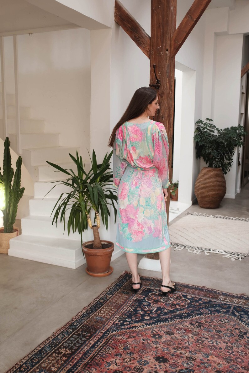 May include: A woman wearing a light blue midi dress with a pink and yellow floral print. The dress has a V-neckline and long sleeves. She is standing on a patterned rug.