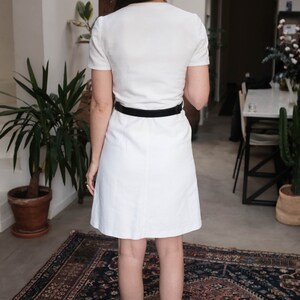 May include: A white dress with short sleeves and a black belt. The dress is knee-length and has a simple, classic design.