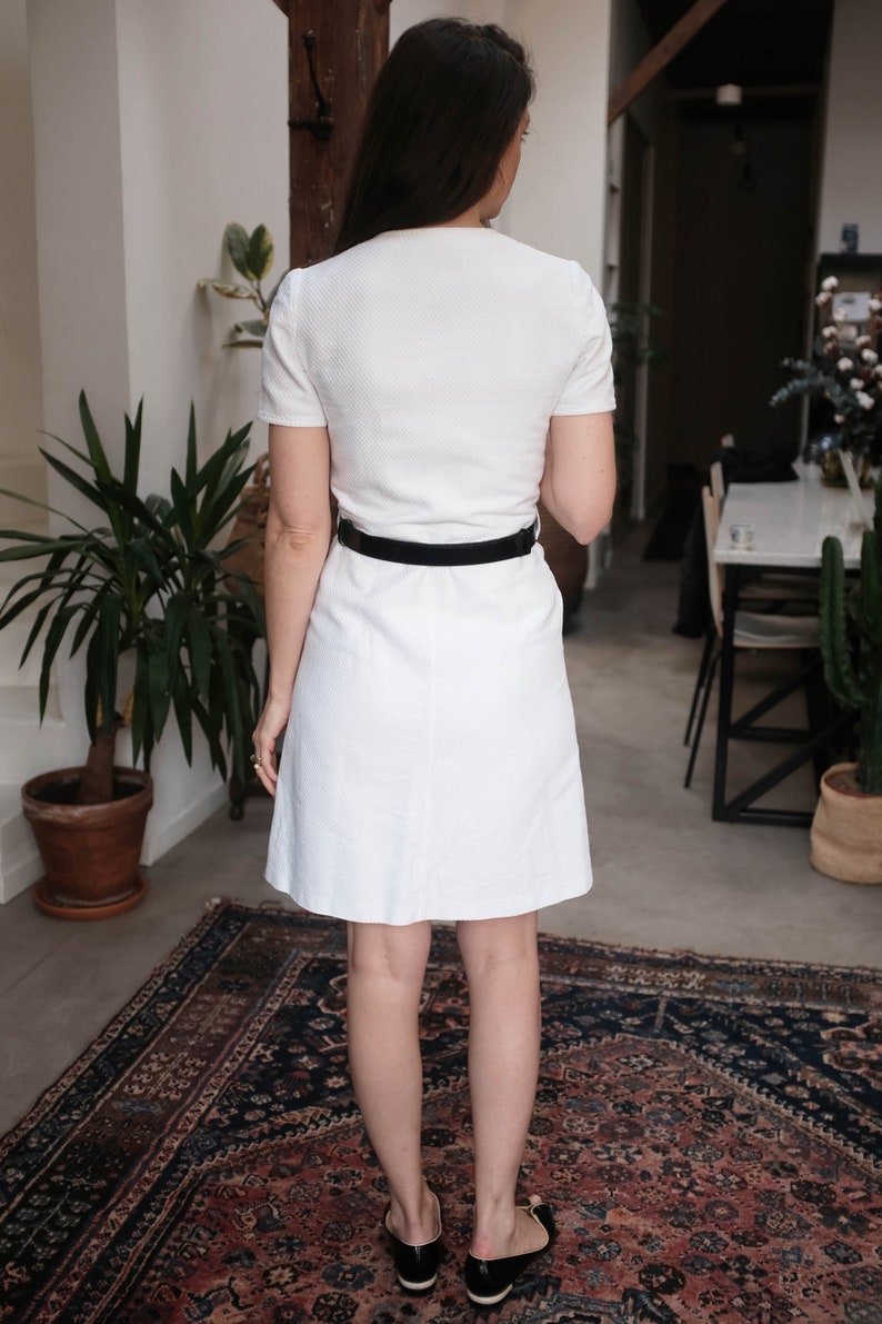 May include: A white dress with short sleeves and a black belt. The dress is knee-length and has a simple, classic design.