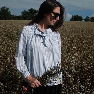 May include: A woman wearing a light blue striped blouse with a bow tie, black sunglasses, and a black skirt, standing in a field of white flowers, holding a bouquet of white flowers.