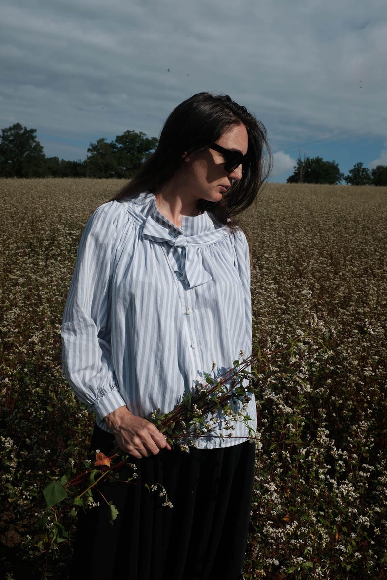 May include: A woman wearing a light blue striped blouse with a bow tie, black sunglasses, and a black skirt, standing in a field of white flowers, holding a bouquet of white flowers.