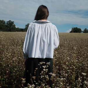 May include: A person wearing a white and blue striped blouse with a gathered neckline and long sleeves, standing in a field of white flowers.