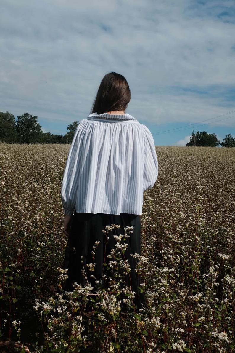 May include: A person wearing a white and blue striped blouse with a gathered neckline and long sleeves, standing in a field of white flowers.