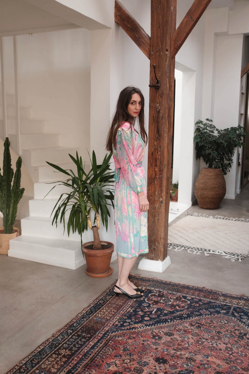 May include: A woman wearing a pink and blue floral print dress with long sleeves and a tie at the waist. She is standing on a patterned rug with a wooden beam behind her.