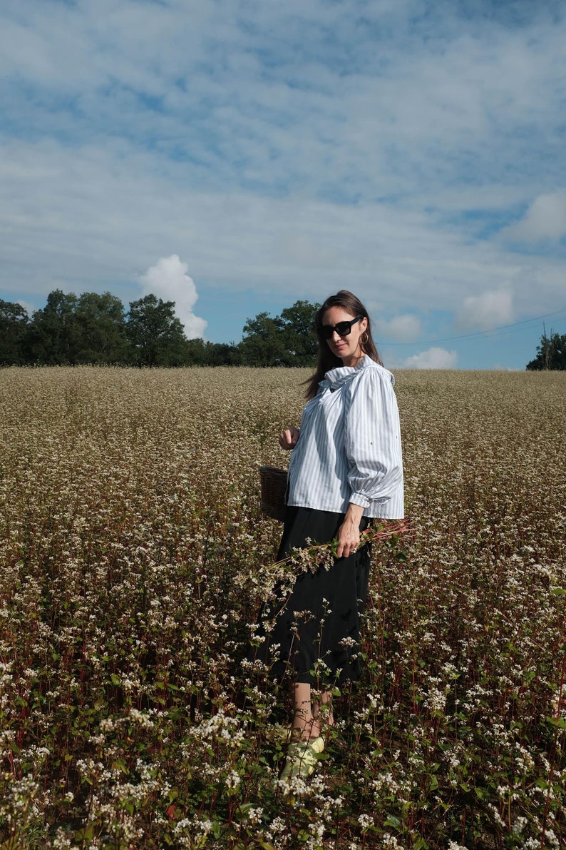 May include: A woman wearing a blue and white striped shirt with a bow tie, black skirt, and sunglasses stands in a field of white buckwheat flowers.