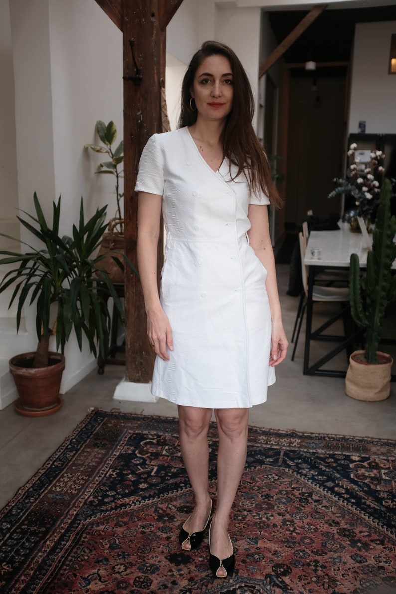 May include: A woman wearing a white, short-sleeved dress with a V-neckline and a double-breasted closure. She is standing on a patterned rug and wearing black shoes.