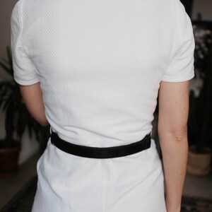 May include: A white dress with a textured fabric and a black belt. The dress has short sleeves and a round neckline.