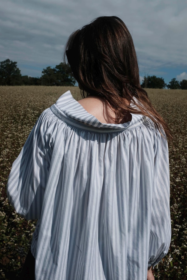 May include: A white and blue striped shirt with a loose, flowing fit and a gathered neckline. The shirt is made of a lightweight fabric and has a relaxed, casual style.