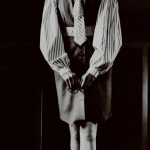 May include: A black and white photo of a person wearing a striped shirt with a tie and a skirt. The person is standing in front of a dark background.
