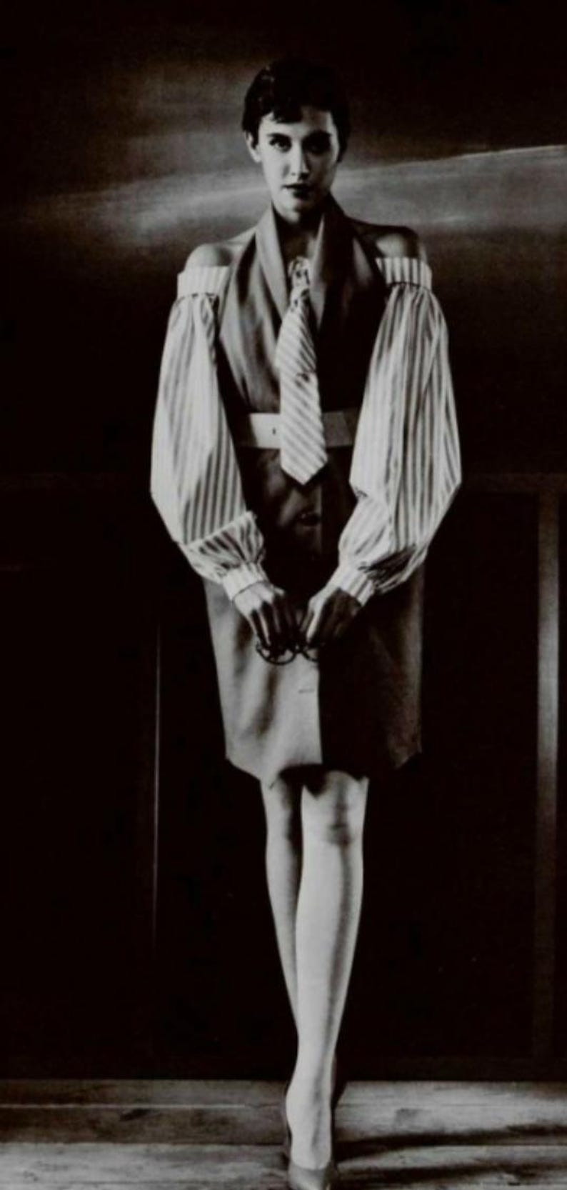 May include: A black and white photo of a person wearing a striped shirt with a tie and a skirt. The person is standing in front of a dark background.