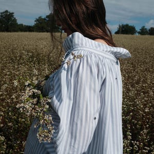 May include: A woman wearing a white and blue striped shirt with a ruffled collar stands in a field of white flowers. The shirt has a relaxed fit and is made of a lightweight fabric.