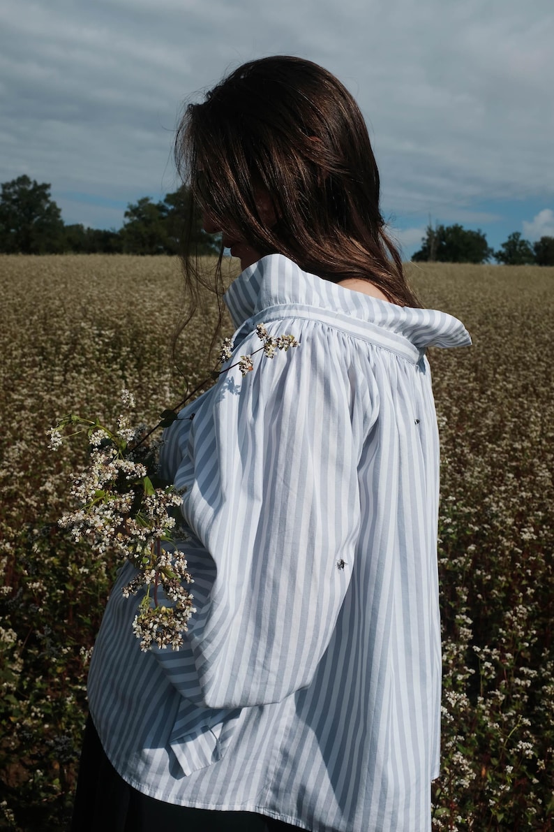 May include: A woman wearing a white and blue striped shirt with a ruffled collar stands in a field of white flowers. The shirt has a relaxed fit and is made of a lightweight fabric.