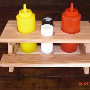 May include: A wooden condiment holder shaped like a miniature picnic table. It holds yellow mustard, red ketchup, and white and black containers. The holder is made of light-colored wood and sits on a dark wooden surface.
