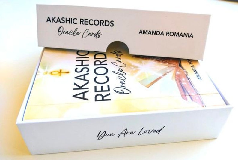 Pu&ograve; includere: Scatola bianca con coperchio bianco contenente un mazzo di carte Oracle Akashic Records. La scatola ha il testo "Akashic Records Oracle Cards" e "You Are Loved" stampato su di essa. Il coperchio ha il testo "Akashic Records Oracle Cards" e "Amanda Romania" stampato su di esso.