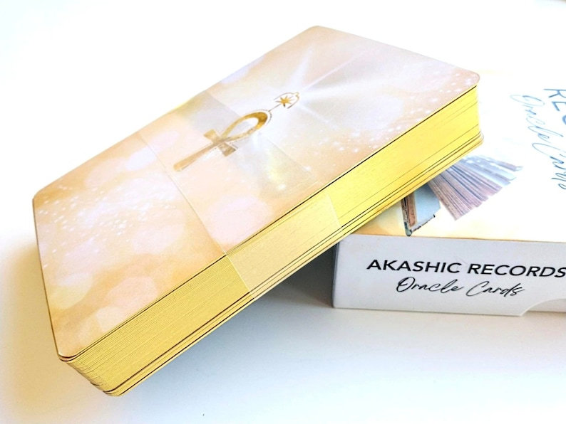 Pu&ograve; includere: Un mazzo di carte Oracle Akashic Records con un design a bordo dorato. La carta superiore presenta un simbolo Ankh dorato su uno sfondo bianco e dorato.