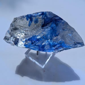 May include: A clear, irregularly shaped glass sculpture with streaks of blue. The artwork is mounted on a clear acrylic stand, casting a shadow below. The piece has a textured surface and appears to be hand-crafted.