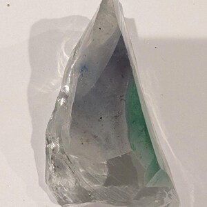 May include: A translucent, irregularly shaped crystal with a sharp point. The crystal has internal colors of green, blue, and gray. The surface is textured and reflects light, creating highlights and shadows.