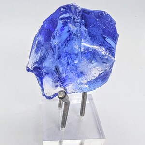 May include: A jagged, translucent cobalt blue glass specimen with visible internal bubbles and textures, mounted on a clear acrylic base with two silver-toned metal prongs that support the piece in an upright, angled position.