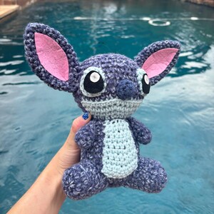 Puede incluir: Mu&ntilde;eco Stitch de ganchillo hecho a mano en tonos azules, grises y rosas. El peluche presenta grandes ojos negros, una barriga azul claro y orejas grandes con detalles interiores rosas. El mu&ntilde;eco est&aacute; sentado con las piernas extendidas.