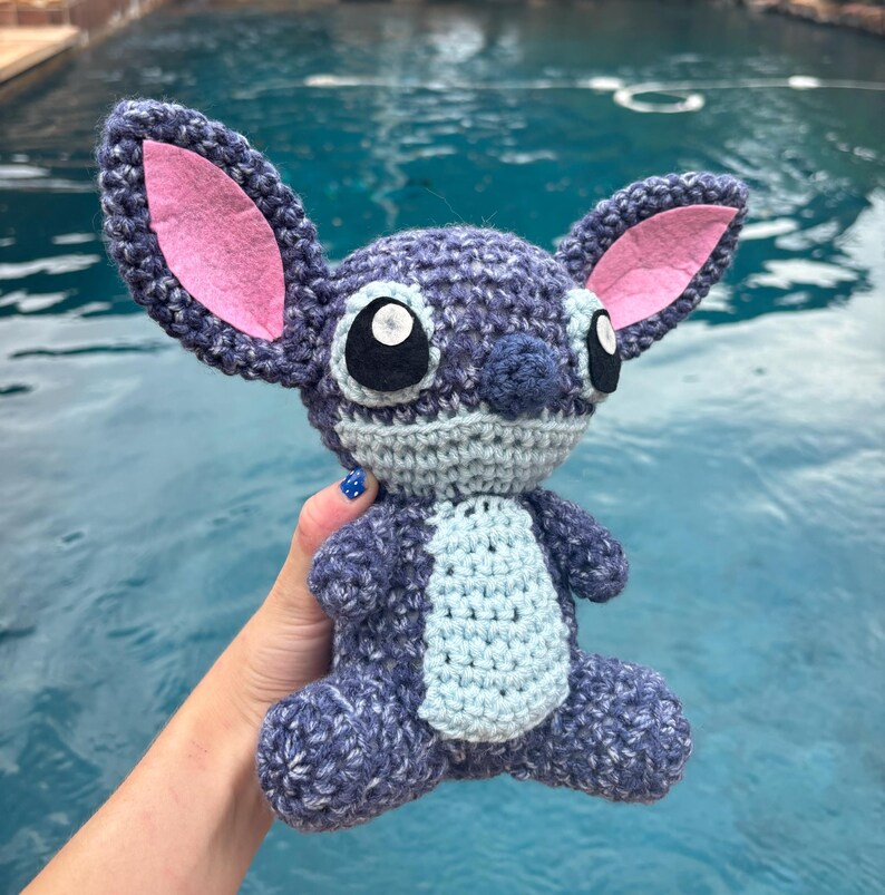Puede incluir: Mu&ntilde;eco Stitch de ganchillo hecho a mano en tonos azules, grises y rosas. El peluche presenta grandes ojos negros, una barriga azul claro y orejas grandes con detalles interiores rosas. El mu&ntilde;eco est&aacute; sentado con las piernas extendidas.
