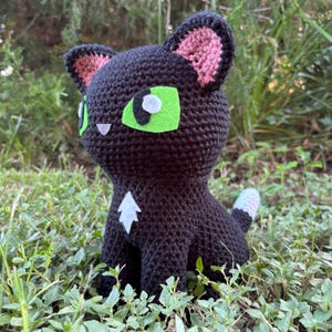 May include: A black crocheted cat plush toy featuring large bright green felt eyes with white pupils, pink inner ears, a small white triangular chest patch, and a white-tipped tail. The yarn texture is visible throughout the entire figure.
