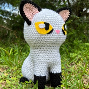 May include: A handmade crochet cat doll in a sitting position. The cat is primarily white with black accents on the paws, ears, and tail. It has yellow eyes, pink inner ears, and a pink nose. The doll is set against a backdrop of green grass.