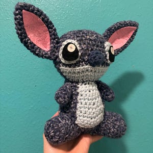 Puede incluir: Un peluche de ganchillo hecho a mano de un personaje alien&iacute;gena azul con grandes orejas y ojos. El juguete tiene una barriga gris y orejas internas rosas. El personaje est&aacute; sentado y parece estar hecho de hilo.