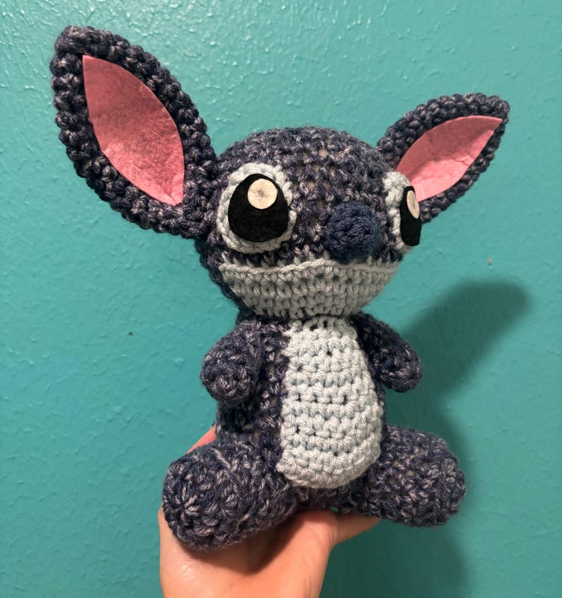 Puede incluir: Un peluche de ganchillo hecho a mano de un personaje alien&iacute;gena azul con grandes orejas y ojos. El juguete tiene una barriga gris y orejas internas rosas. El personaje est&aacute; sentado y parece estar hecho de hilo.