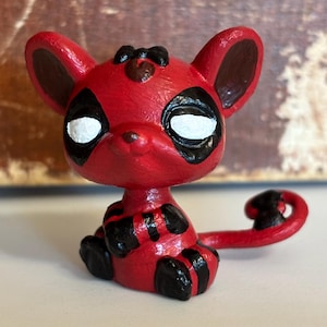 Puede incluir: Una figura roja de un animal de dibujos animados con marcas negras alrededor de los ojos, en las orejas y en el cuerpo. Los ojos son blancos. La cola está enroscada. La figura está sobre una superficie blanca.