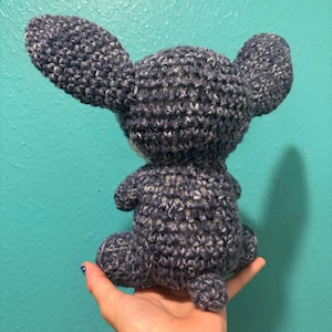 Puede incluir: Un peluche de ganchillo hecho a mano, visto desde atr&aacute;s, en posici&oacute;n sentada. El juguete es de color azul oscuro con motas blancas, con orejas grandes y una cola peque&ntilde;a. El juguete se sostiene en una mano.